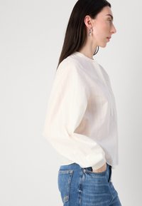 Una donna con lunghi capelli scuri posa di profilo, indossando una blusa bianca a righe e jeans blu, con uno sfondo minimalista.