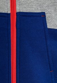 Hoodie zippé bleu et gris avec une fermeture éclair rouge, texture de tissu lisse, et design de panneaux contrastants avec des poches latérales.