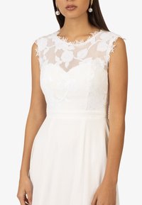 Weißes Kleid mit einem floralen Spitzenoberteil und transparenter Überlagerung, mit rundem Ausschnitt und weichem, fließendem Rock. Elegante Details an den Schultern.
