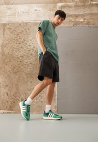 Grön bomullströja, svarta denimshorts och gröna sneakers med vita ränder. Bakgrunden har texturerade betongväggar och golv.