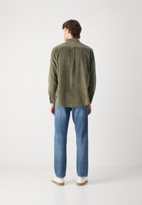 Olijfgroene corduroy shirt met lange mouwen, detail van een rugplooi en een ronde zoom. Gecombineerd met lichtblauwe denim jeans en witte schoenen.