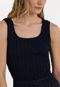 Débardeur tricoté bleu marine avec un décolleté carré et une texture en côtes mettant en avant des motifs de tricot en relief. Design sans manches avec une silhouette ajustée.