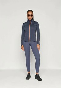 Kék teljesítménydzseki narancssárga cipzárral, hozzá illő leggingszel párosítva. Simasága és testhezálló fazonja kiemelkedő, fekete sportcipővel kiegészítve.