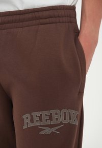 Pantalones de chándal marrones con una textura suave y cintura elástica. Presenta un logo gris de "REEBOK" y bolsillos laterales para mayor practicidad.