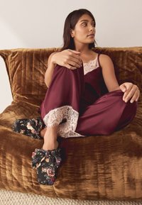 Conjunto de pijama de satén burdeos con ribete de encaje; zapatillas con nudo y estampado floral; sobre un cojín marrón con textura.