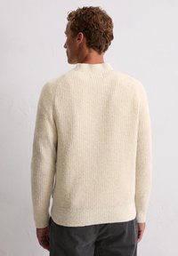 Cremefarbener gerippter Pullover mit hohem Kragen und langen Ärmeln. Weiche Textur mit dezenten Farbnuancen. Der Schnitt ist leicht oversized.