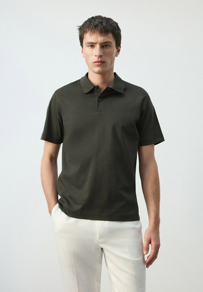 Joven de pie con las manos en los bolsillos, vistiendo una camiseta polo verde oscuro y pantalones blancos contra un fondo claro y liso.