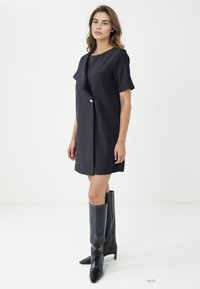 Vestito nero a maniche corte con pannello anteriore svasato, impreziosito da un dettaglio con bottone dorato. Abbinato a stivali neri in pelle al ginocchio.