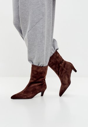 Bottines en suede marron avec un bout pointu et un talon haut élégant, portées avec un pantalon de survêtement gris, mettant en avant une texture lisse.