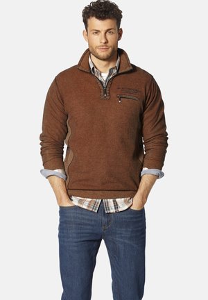 Brauner Fleece-Pullover mit Viertelreißverschluss, strukturiertem Stoff und schwarzem Reißverschluss. Über einem karierten Hemd getragen. Blaue Jeans runden das Outfit ab.