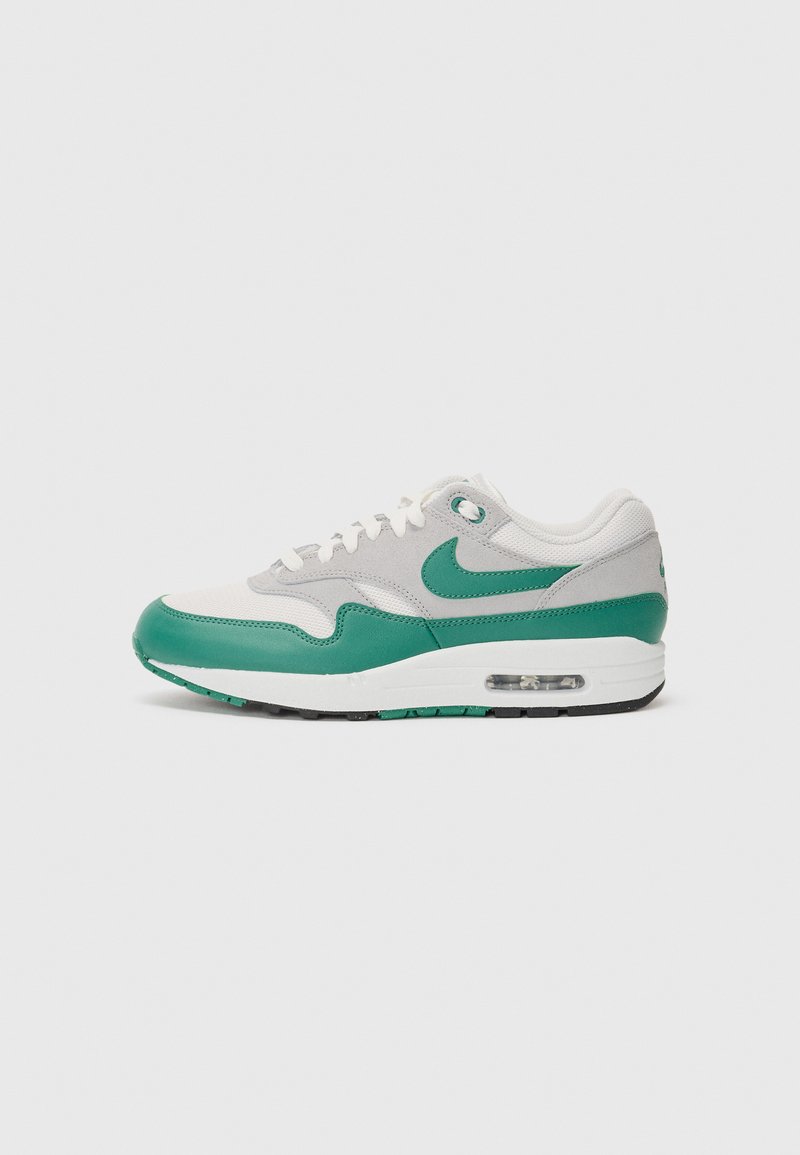 Nike Air Max 1 športni čevlji z zgornjim delom iz sive mrežice, zelenimi usnjenimi detajli, belimi vezalkami in vidno zračno enoto v podplatu.