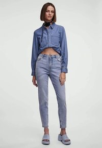 Svetlomodrá denimová skrátená košeľa s gombíkmi vpredu a dvomi náprsníkmi, skombinovaná so svetlomodrými vysoko pásovými džínami a svetlomodrými sandálmi na podpätku.