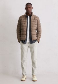 Beige donsjack met gewatteerd ontwerp, ritssluiting en opstaande kraag. Gecombineerd met een grijs overhemd met knopen, witte T-shirt, lichte broek en sneakers.