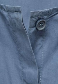 Gray button-up shirt featuring a collar, smooth texture, and side stitching detail. The button is a round gray plastic with four holes.