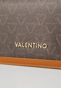 Brun läderväska med struktur och en guldfärgad "VALENTINO"-logotyp, med en beige accentremsa längs botten. Mönstrad med V-former.