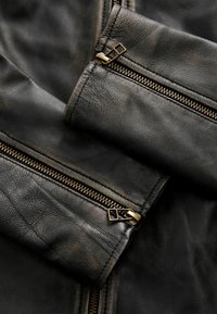 Zwarte leren jas met een gestructureerde afwerking, voorzien van gouden ritsen en hardware. Close-up van de details van de mouwen en de voorrits.