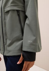 Veste softshell verte avec des accents noirs, dotée de poches zippées et du logo "Cecil Outerwear" sur la manche. Texture de tissu lisse.