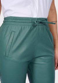 Pantalones jogger de cuero sintético en color verde azulado con cintura elástica, cordón y un bolsillo lateral, que presentan una textura suave y mate.