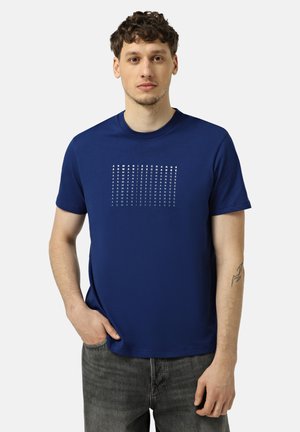 Uomo che indossa una t-shirt blu navy Armani Exchange con stampa ripetuta del nome del brand, jeans grigio scuro e tatuaggio al braccio, in piedi con una mano in tasca.