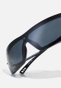 Lunettes de soleil noires avec de grandes montures aérodynamiques et des verres teintés foncés. Parmi les caractéristiques figurent un design élégant et des branches ajustables avec des détails de taille.