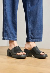 Mule aperte in pelle nera con punta aperta, dotate di un tacco curvo e spesso e di una suola grigia testurizzata, indossate con pantaloni in denim scuro su un pavimento di legno.