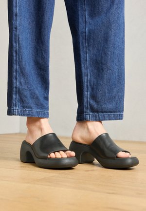 Czarne skórzane mules z odkrytymi palcami, z zakrzywionym, masywnym obcasem i teksturowaną szaro-skalistą podeszwą, noszone z ciemnoniebieskimi spodniami dżinsowymi na drewnianej podłodze.