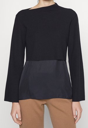 Personne portant un haut noir à manches longues superposé avec un col bateau, sur une sous-couche noire ample, et un pantalon de couleurcamel.