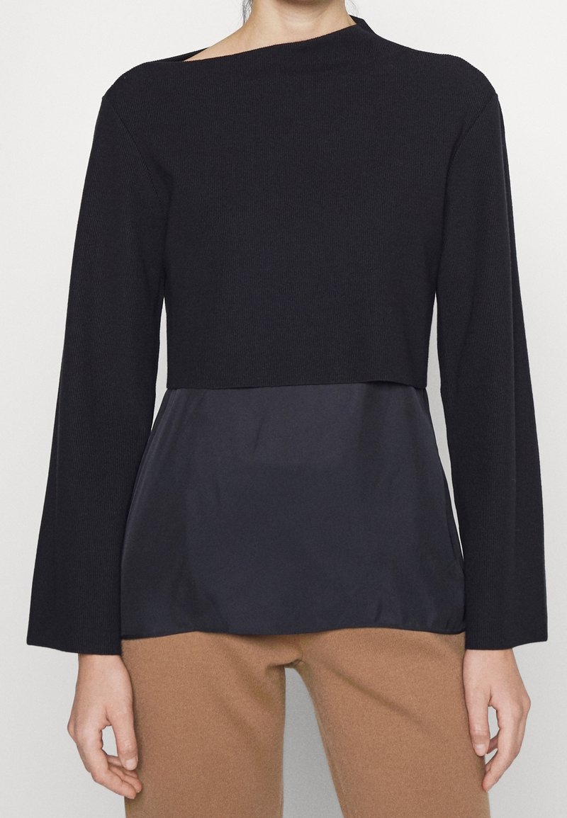 Osoba nosząca czarną, warstwową bluzkę z długim rękawem i dekoltem typu łódka, na luźnej, czarnej warstwie pod spodem oraz camelowe spodnie.