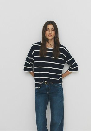 Moteris, turinti ilgus rudus plaukus, dėvi juodai baltą dryžuotą marškinėlį ir mėlynas džinsines kelnes, stovinti rankomis už nugaros prie baltos sienos.