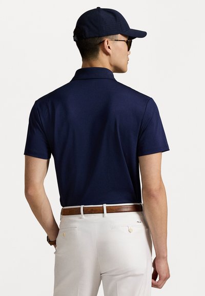 Polo cu guler de culoare bleumarin, asortat cu pantaloni scurți de culoare deschisă, croiți pe corp, și o curea din piele maro, prezentat din spate.