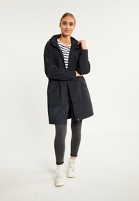 Lange schwarze wasserdichte Jacke mit Reißverschluss, Kapuze und zwei Fronttaschen, getragen über einem gestreiften Hemd und grauen schmal geschnittenen Hosen.