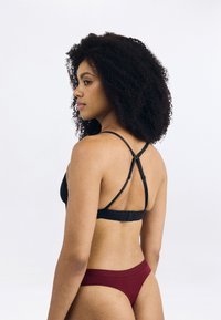 Črna čipkast bralette s prilagodljivimi trakovi in zapenjanjem na hrbtu, v kombinaciji z burgundskimi hipster hlačkami. Gladka tekstura, minimalističen dizajn.