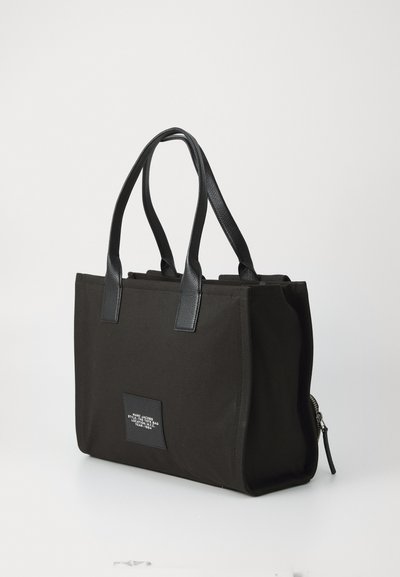 Geantă tote neagră, rectangulară, cu două mânere din piele și un eticheta frontală care afișează text alb de la Marc Jacobs.