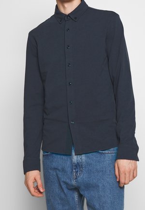 Marineblauwe overhemd met lange mouwen en knoopsluiting, voorzien van een puntige kraag, donkere knopen, een slim fit en een zachte textuur. Gekocht in combinatie met een blauwe spijkerbroek.