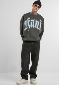 Felpa oversized grigia scura con grafica "kani", abbinata a jeans neri larghi e sneakers crema. Il modello indossa un berretto nero e occhiali.