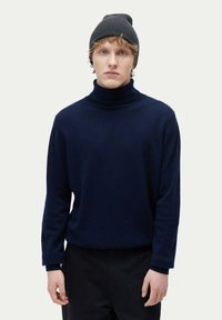 Marinblå turtleneck-tröja i strukturerad stickad kvalitet, som är kombinerad med en mörkgrå ribbad mössa. Modellen står mot en ljus bakgrund.