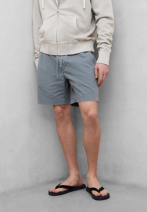 Mann iført lysegrå shorts, beige hettejakke med glidelås og svarte flip-flops, stående mot en ensfarget lysegrå bakgrunn.