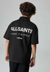 Vaikas stovi nugarą į kamerą, vilkintis juodą marškinėlį su užrašu "ALLSAINTS LONDON" ir papildomais japonų simboliais.