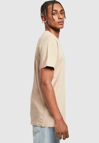 Beige bomulds t-shirt med korte ærmer, rund halsudskæring og afslappet pasform, vist af en model fra siden mod en neutral baggrund.