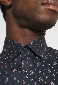 Marinblå skjorta med knappar och ett litet blommönster i rött och grönt. Har en vanlig krage och texturerad tyg.