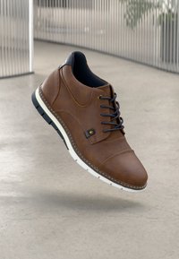 Scarpa marrone in pelle con allacciatura, dalla texture liscia, dotata di suola in gomma bianca e interno rivestito in nero. Presenta un piccolo dettaglio in metallo.