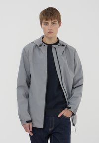 Giacca a hood grigia chiara con zip frontale, regolabile con lacci e polsini elastici. Indossata sopra un maglione blu scuro e pantaloni di denim.