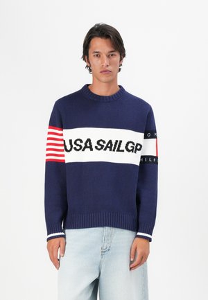 Navy blue knitted sweater with white and red horizontal stripes featuring "USA SAIL GP" text. Ribbed collar and cuffs, casual fit.