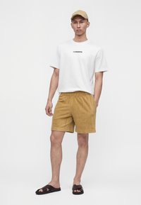 Homme portant une casquette beige, un t-shirt blanc J.Lindeberg, un short en tricot moutarde et des sandales slides marron foncé, debout sur fond blanc.