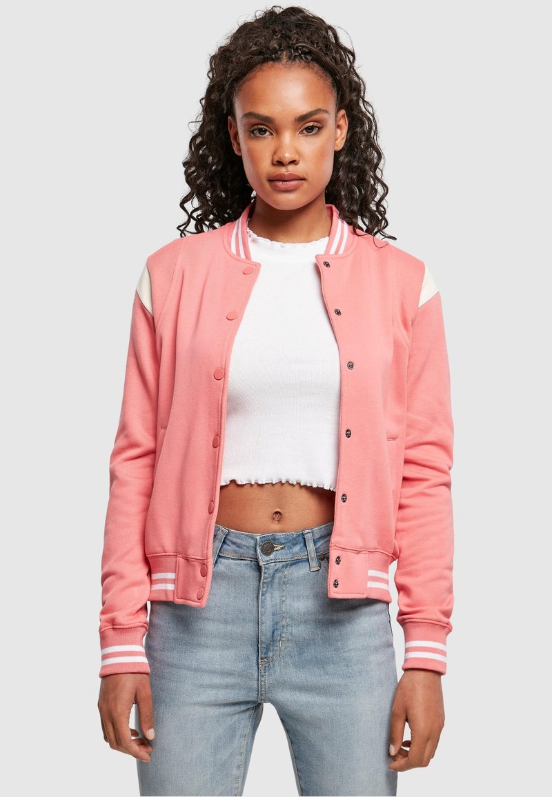 Roze bomberjack met witte accenten, drukknoopsluiting, geribbelde manchetten en zoom, gedragen over een wit cropped topje, gecombineerd met lichte jeans.