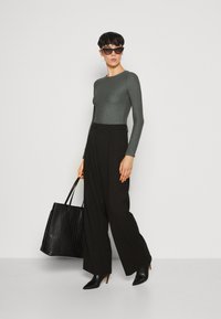Graues, strukturiertes Langarmoberteil, weite schwarze Hose und eine schwarze gefaltete Tragetasche. Abgerundet mit spitzen schwarzen Pumps und Cat-Eye-Sonnenbrille.