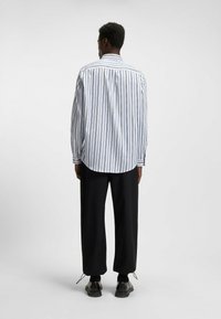 Camicia da uomo con bottoni, a righe verticali blu e bianche, maniche lunghe, orlo arrotondato, indossata con pantaloni neri larghi e scarpe.