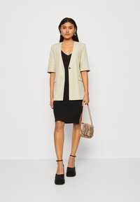 Beige kortärmad blazer med en enkel knapp, över en svart figurnära klänning. Modellen håller en liten, mönstrad väska och bär svarta platåskor.