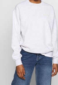 Světlě šedá mikina s dlouhými rukávy a žebrovanými manžetami. Má malý logo značky v neonově žluté barvě. Kombinováno s modrými džínami.