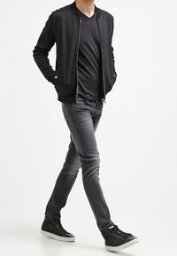 Svart bomberjacka över en svart T-shirt, parat med slim-fit grå jeans och mörka sneakers. Enkel design, mjuk textur och modern passform.